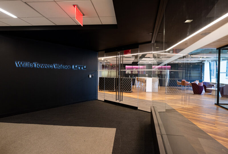 The entrance way to a unit in a large building. Glass door and panels to the right and a backlit sign to the left of the entrance door.