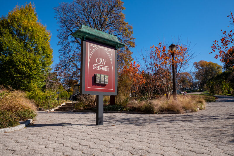 A sign saying Greenwood with brochure holders attached on a cement block lined path with trees in the background