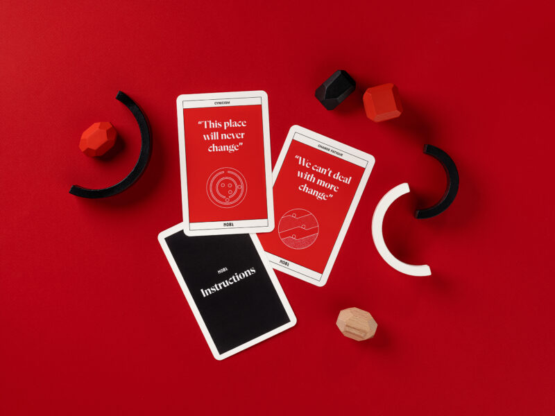 An overhead view of 3 cards: A black instruction card, and two red change barrier cards that read: 'This place will never change', and 'We can't deal with more change'.