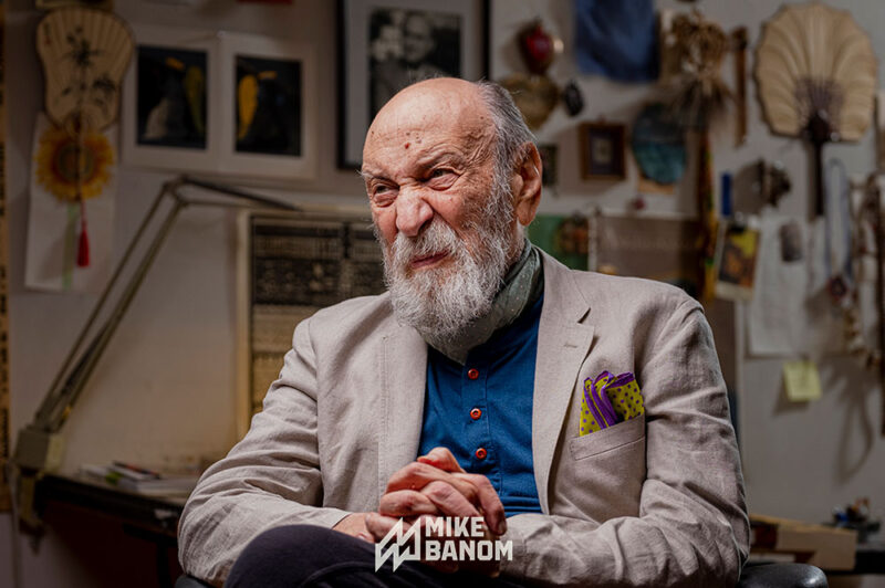 A portrait of a elder man, a legendary graphic designer who is sitting in a chair with his legs crossed and hands clasped. He is in his office surrounded by inspiration hung on the wall behind him.