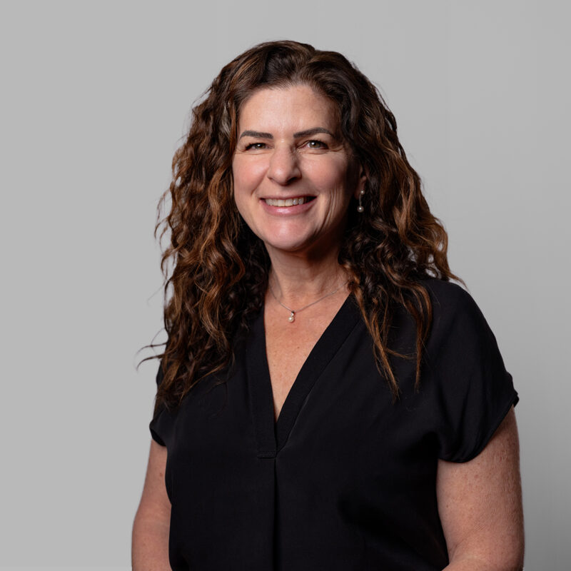 A portrait of a woman smiling in a black outfit and brown curly hair on a grey background