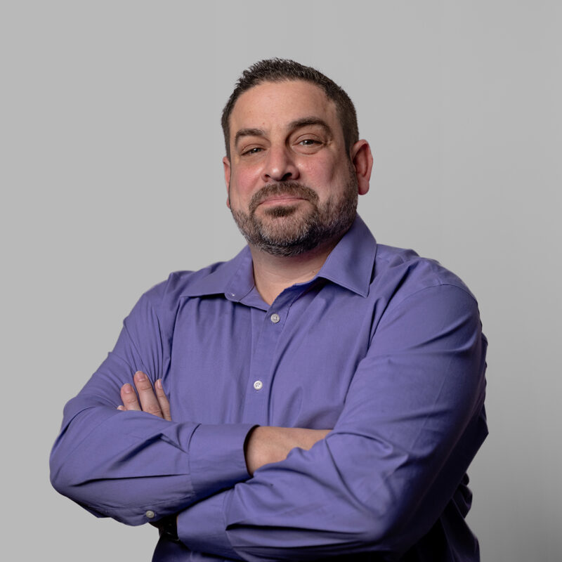 A portrait of a man smiling in a purple shirt and his arms crossed on a grey background