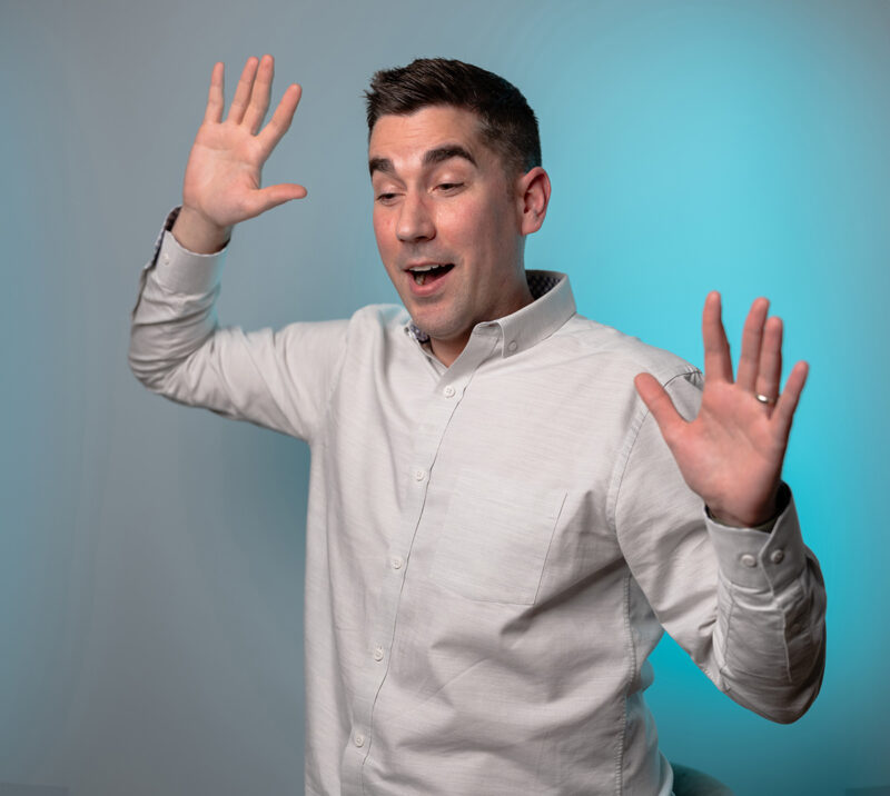 A portrait of a man holding up his hands with a shocked expression on his face featuring a blue LED light filling in the right side of the grey background