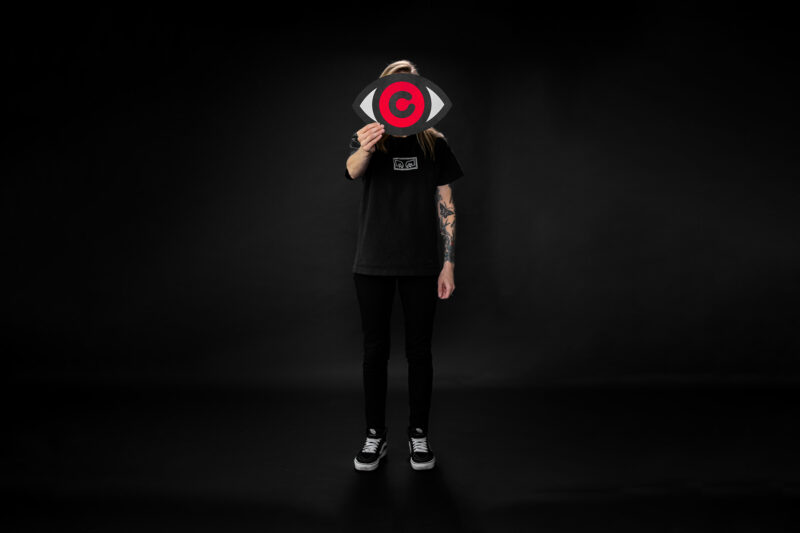 A full body portrait of a woman on an all black background holding up her brand's logo - an eyeball with a red iris and the letter C as the pupil in the middle.