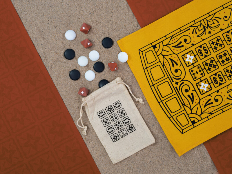 Pieces of a board game lying on sand and red paper including dice, round markers, a material board and a cloth bag.