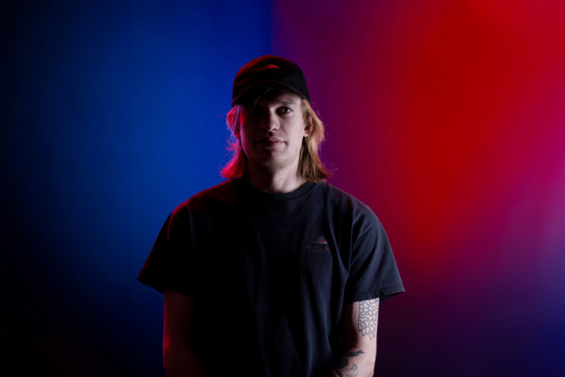 A portrait of a man wearing a hat on a background split down the middle with the left side being blue, and the right side being red. He has a black shirt on and has blond hair.