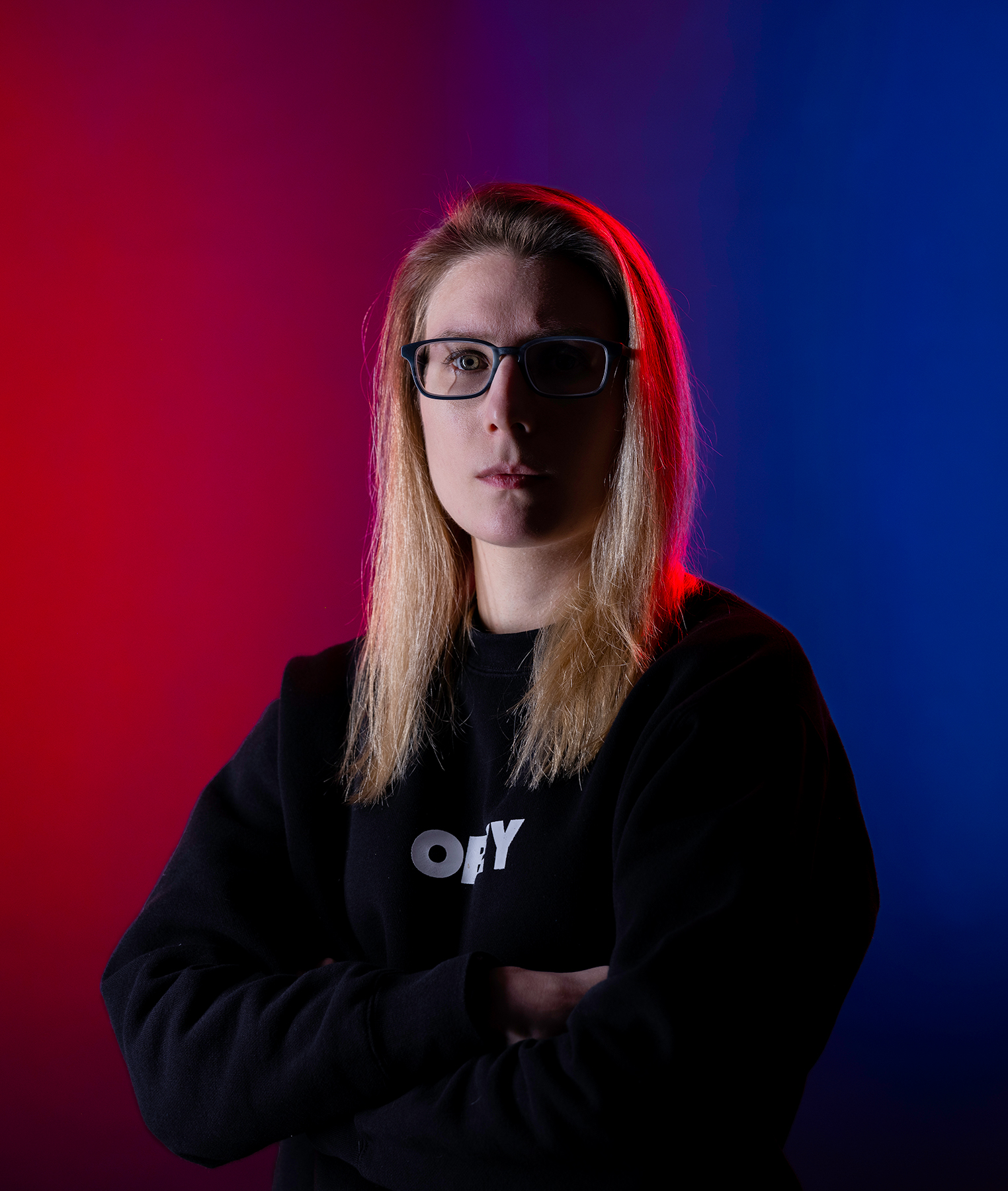 A portrait of a woman crossing her arms in a black hoodie. The background is colored and split down the middle with blue on the right side and red on the left. There is also a highlight of red light shining on her right side contrasting the blue background.