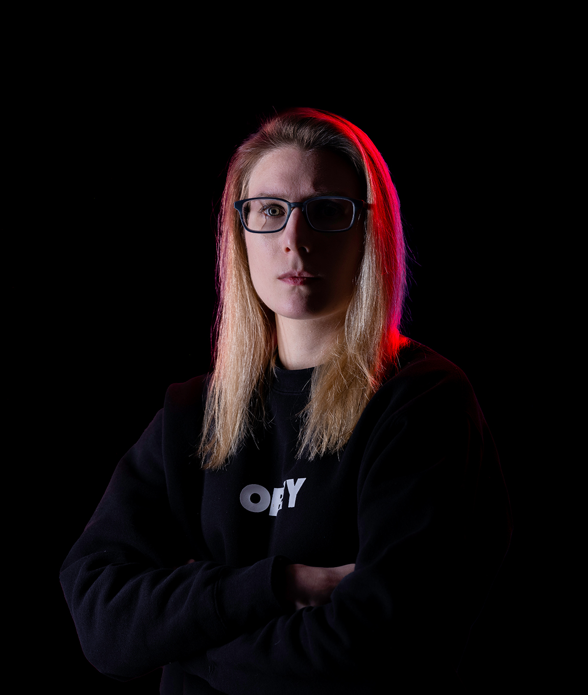 A portrait of a woman crossing her arms in a black hoodie. The background is black. There is also a highlight of red light shining on her right side.