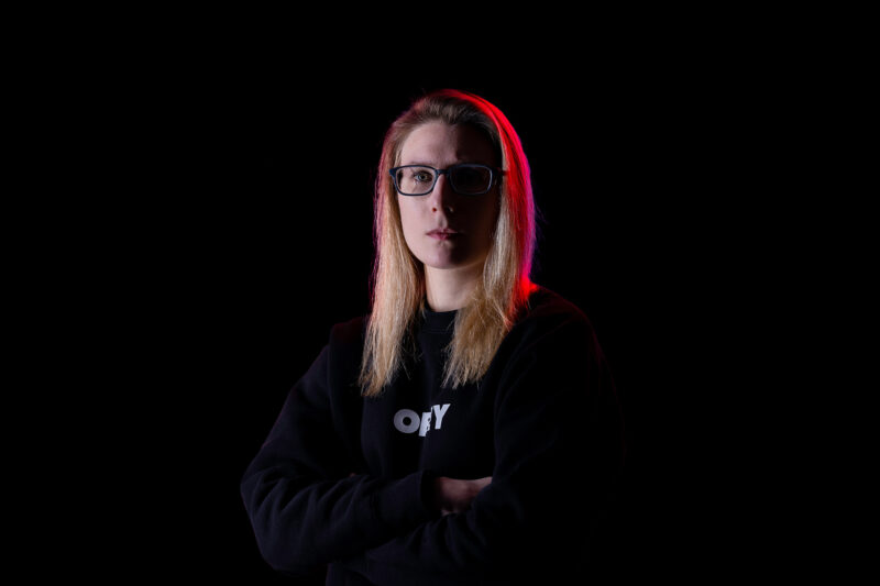 A portrait of a woman with glasses and blonde hair on a black background with a red light highlight on her hair.