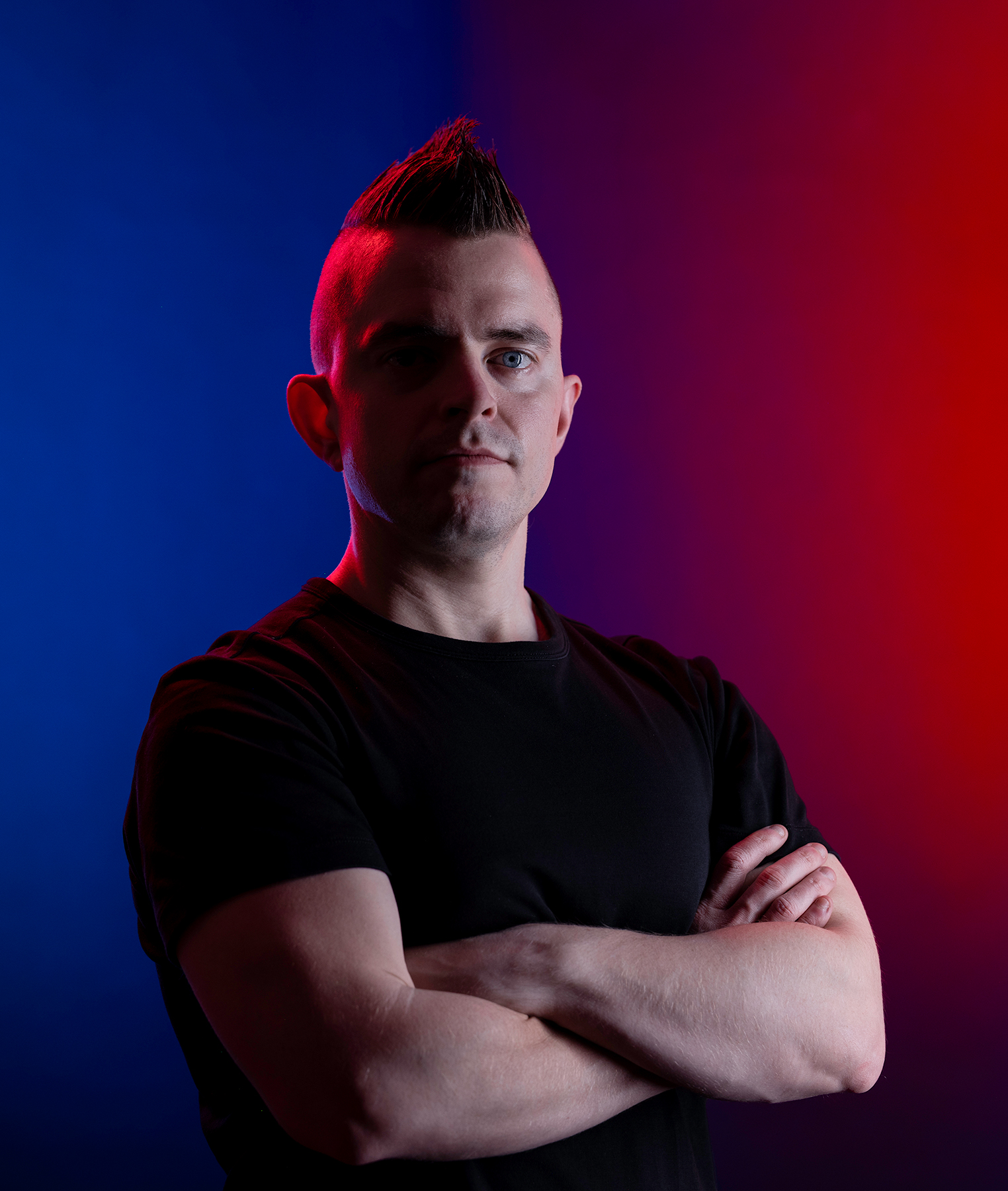 A portrait of a man crossing his arms in a black t-shirt. The background is colored and split down the middle with blue on the left side and red on the right. There is also a highlight of red light shining on his left side contrasting the blue background.