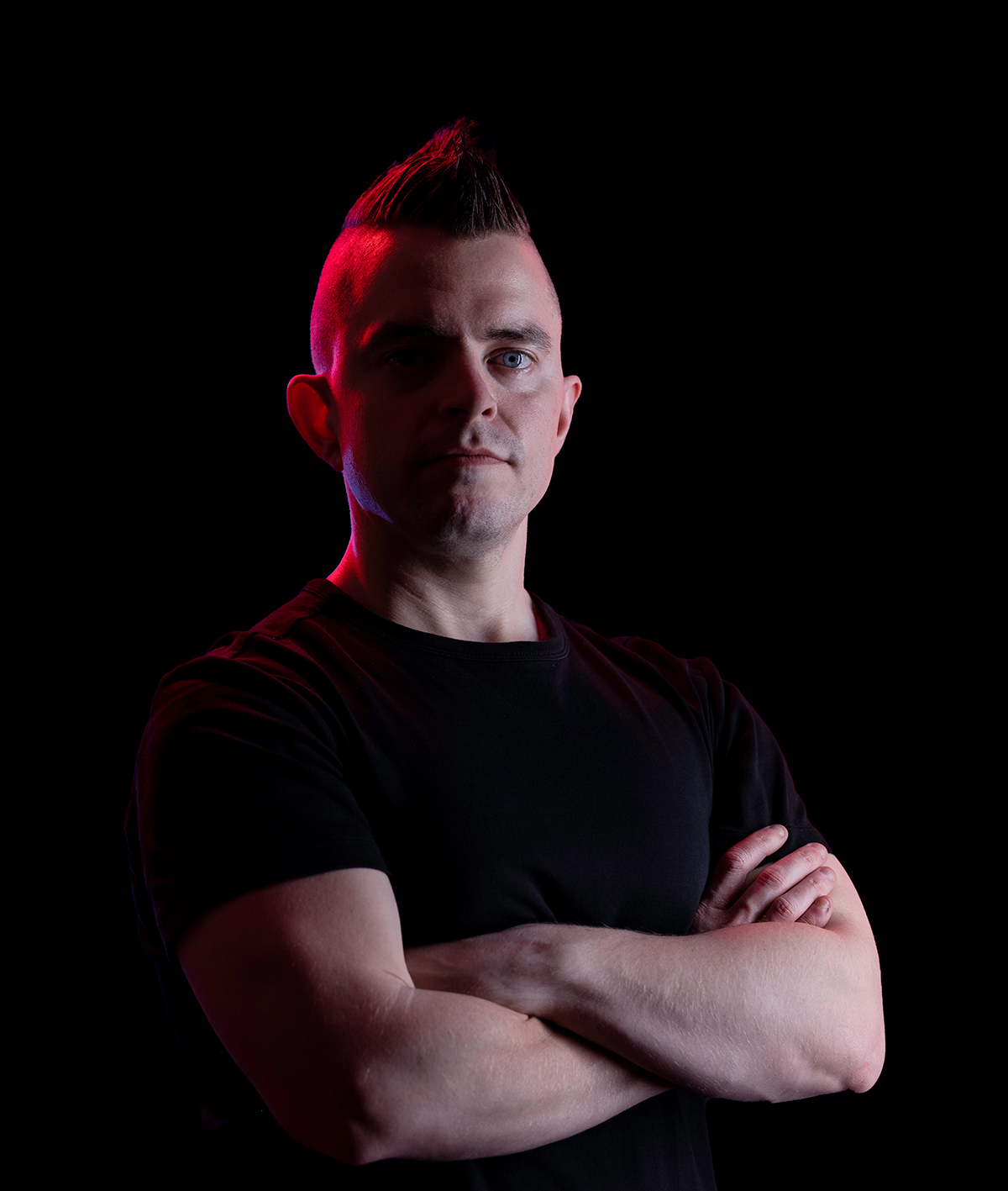 A portrait of a man crossing his arms in a black t-shirt. The background is black. There is also a highlight of red light shining on his left side.