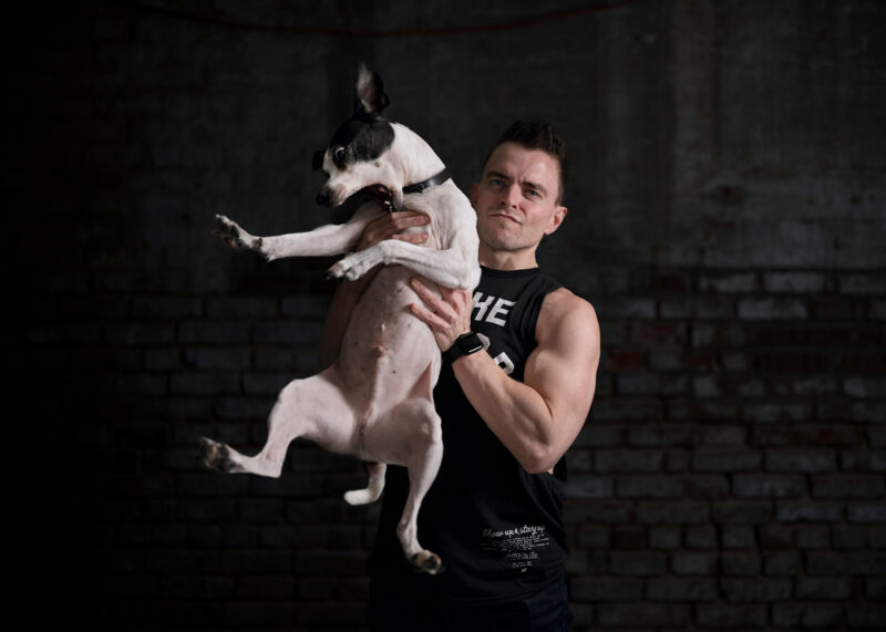 A portrait of a man with muscles in a tank top holding up his black and white terrier dog who is trying get down. The portrait is moody and features a dark brick wall in the background.