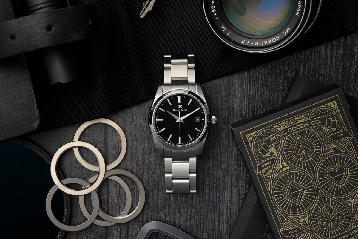 A black dial, silver steel bracelet Grand Seiko watch centered surrounded by several objects on a wood grain background. A vintage minolta lens, porsche key, deck of playing cards, carabiner, and flash light surround the watch. The image is moodily lit.