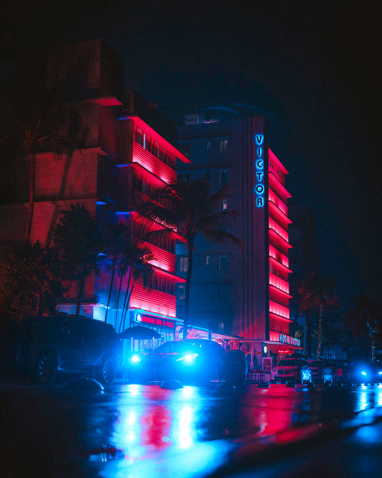 A scene from Miami beach - a car driving down the street with teal headlight beams and a pink illuminated building in the background.