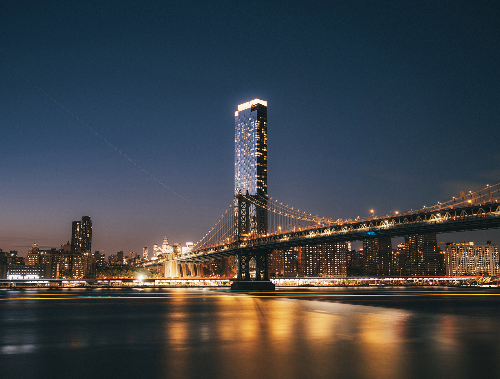 The manhattan bridge at night captured via a long exposure showing a very smooth water with yellow lights