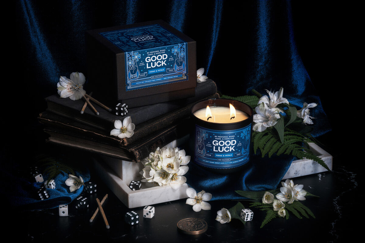 A dramatically lit scene with old books, white marble, white flowers, ferns, and dice featuring a scented candle with an illustrated blue label that reads Good Luck.