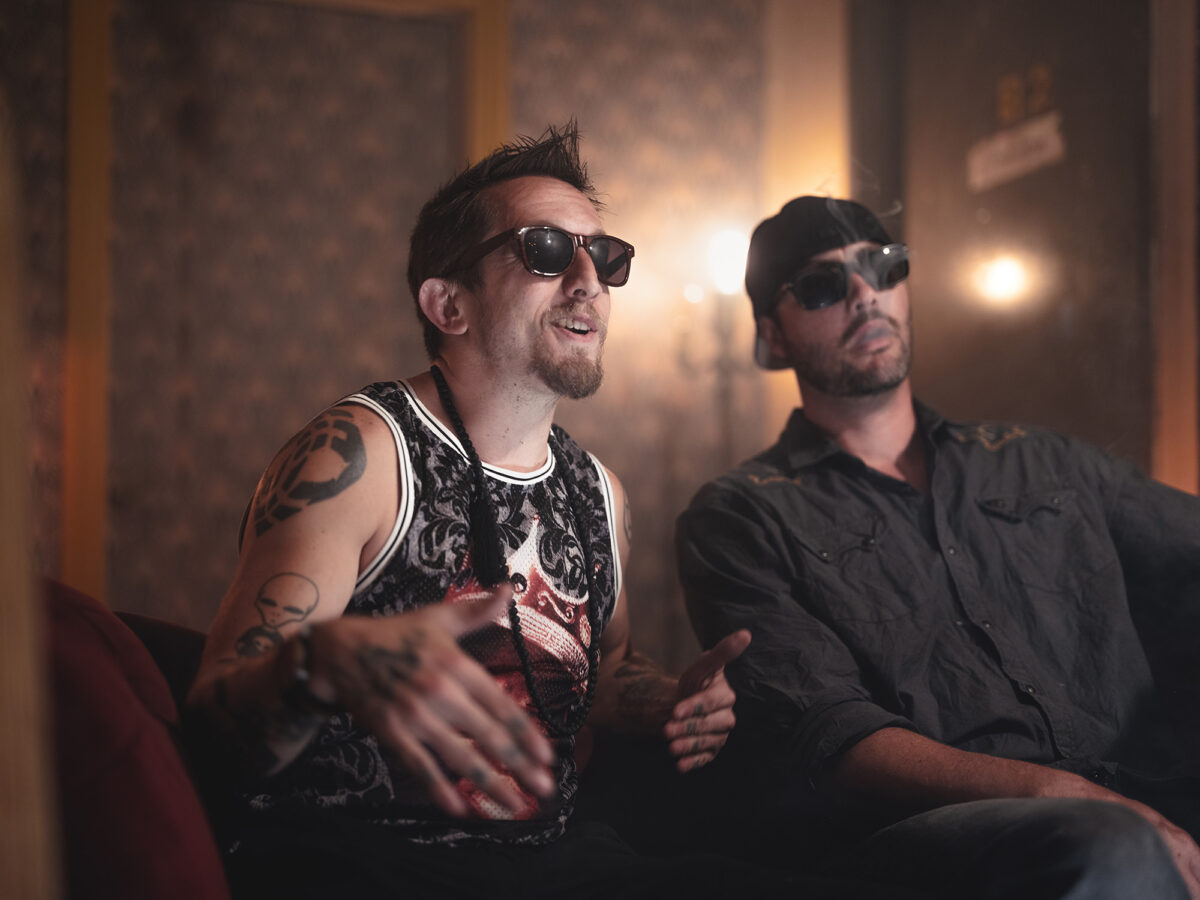 The tattooed man and the man in the button-up shirt sit on a red couch, both wearing dark sunglasses. They appear mid-conversation, relaxed but expressive.
