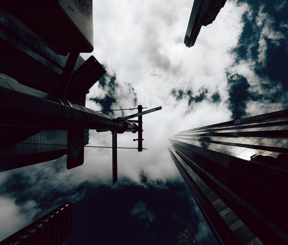 Black-and-white view looking up at a traffic light and towering skyscrapers, with a cloudy sky reflected on the glass.