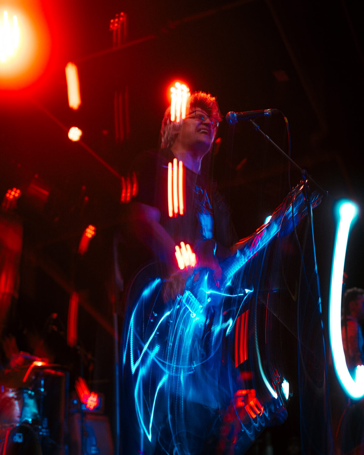 Punk rock guitarist plays electric guitar with red and blue light streaks creating motion blur effect on stage.