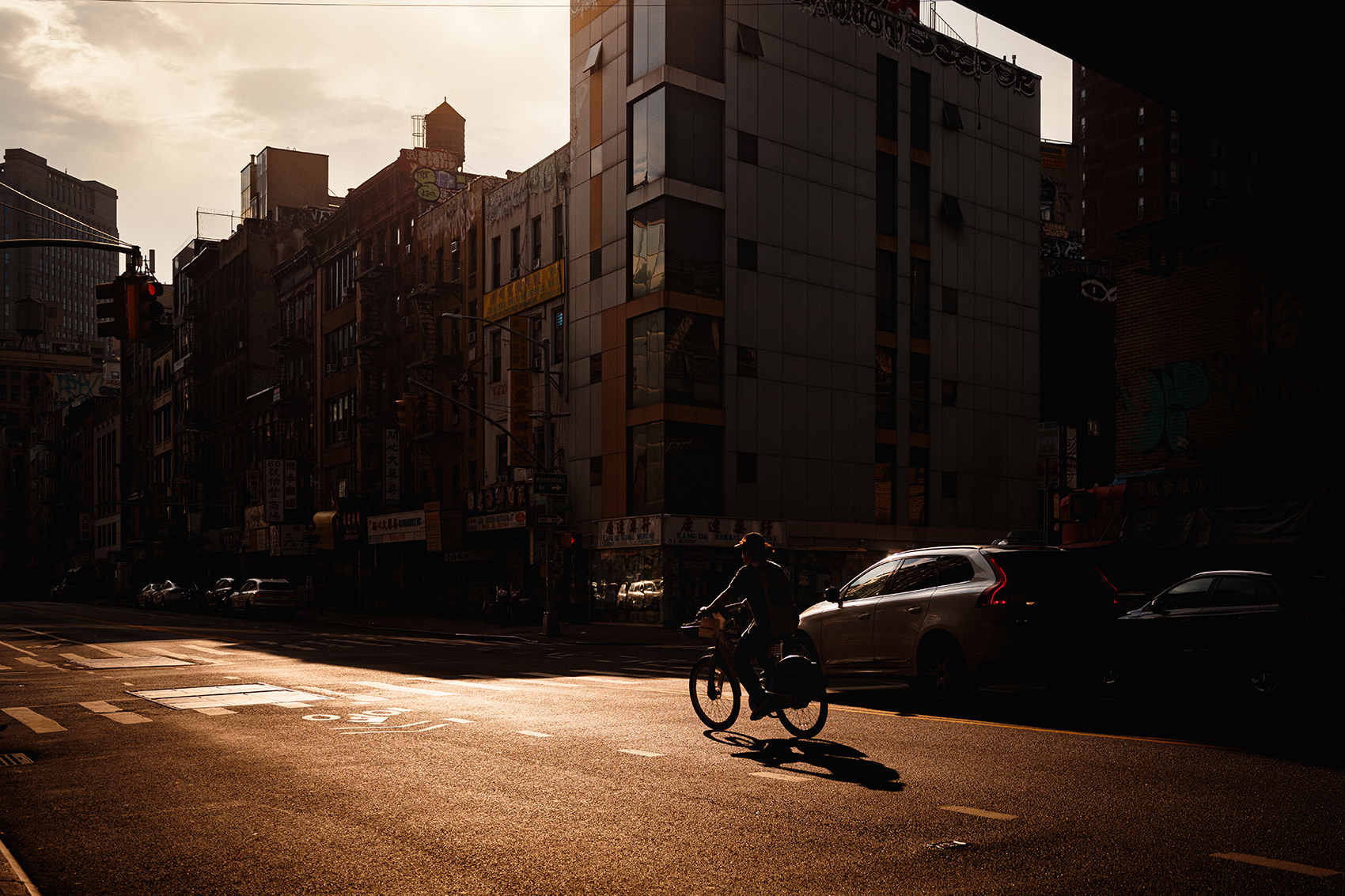 Sunlit Chinatown, NYC intersection with cyclist and parked cars beside a building glowing in late-afternoon light