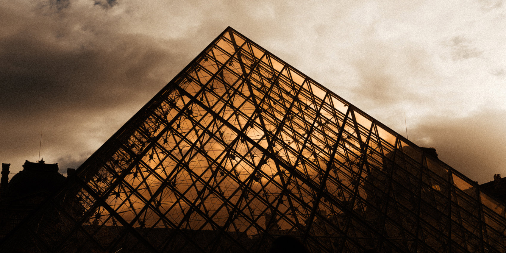 Dramatic low-angle photograph of the Louvre's glass pyramid in Paris, with illuminated golden glass panels, dark geometric framework, and textured clouds overhead.