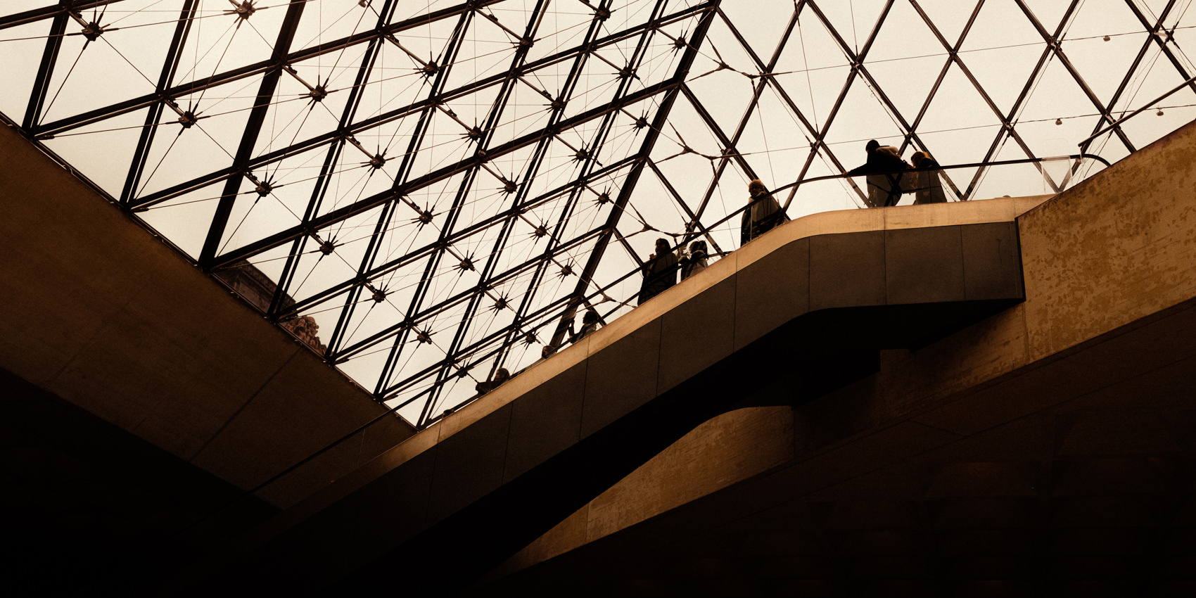 Visitors appear as small silhouettes along a diagonal walkway beneath the Louvre's glass pyramid, surrounded by repeating triangular steel framing and warm natural light.