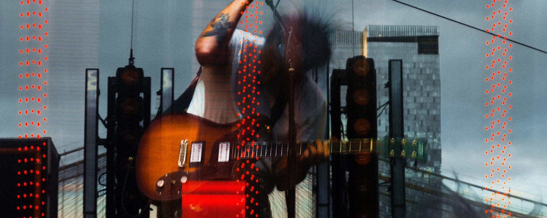 Experimental double-exposure photograph of the Teenage Mortgage guitarist performing with red stage lights and the Brooklyn skyline behind him.
