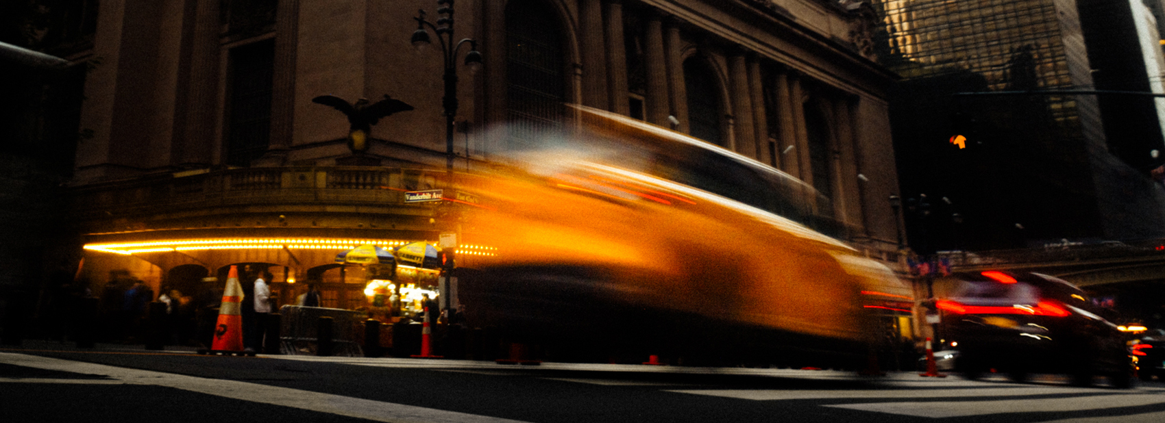 A yellow taxi streaks past Grand Central Terminal in a long-exposure blur, with lights glowing under the entrance canopy.